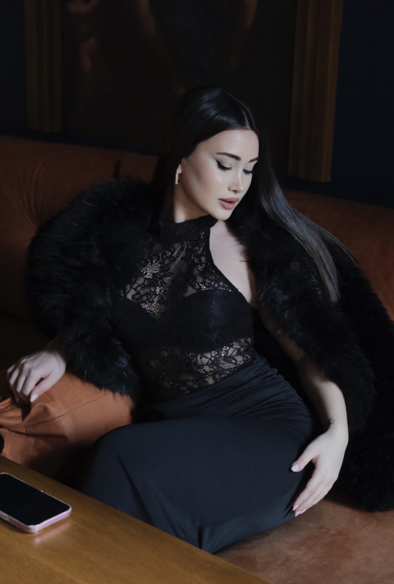 Elegant woman in a black lace top and fur wrap sits on a brown sofa, looking downward to her left.