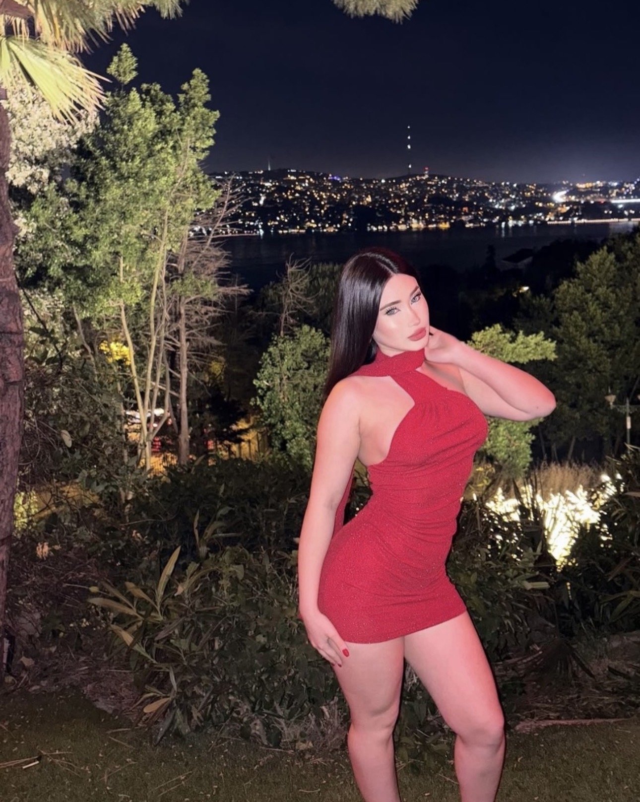 Woman in a red halter-neck bodycon dress posing in a garden at night with city lights in the distance behind her.