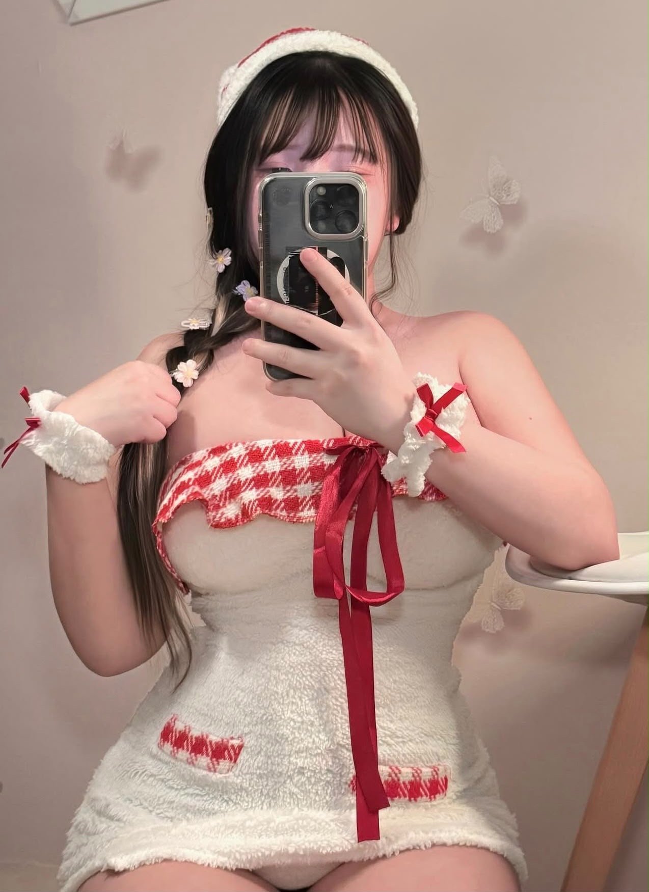 Woman in a fluffy white towel dress with red plaid trim, braided hair with flower clips, taking a mirror selfie with a phone.