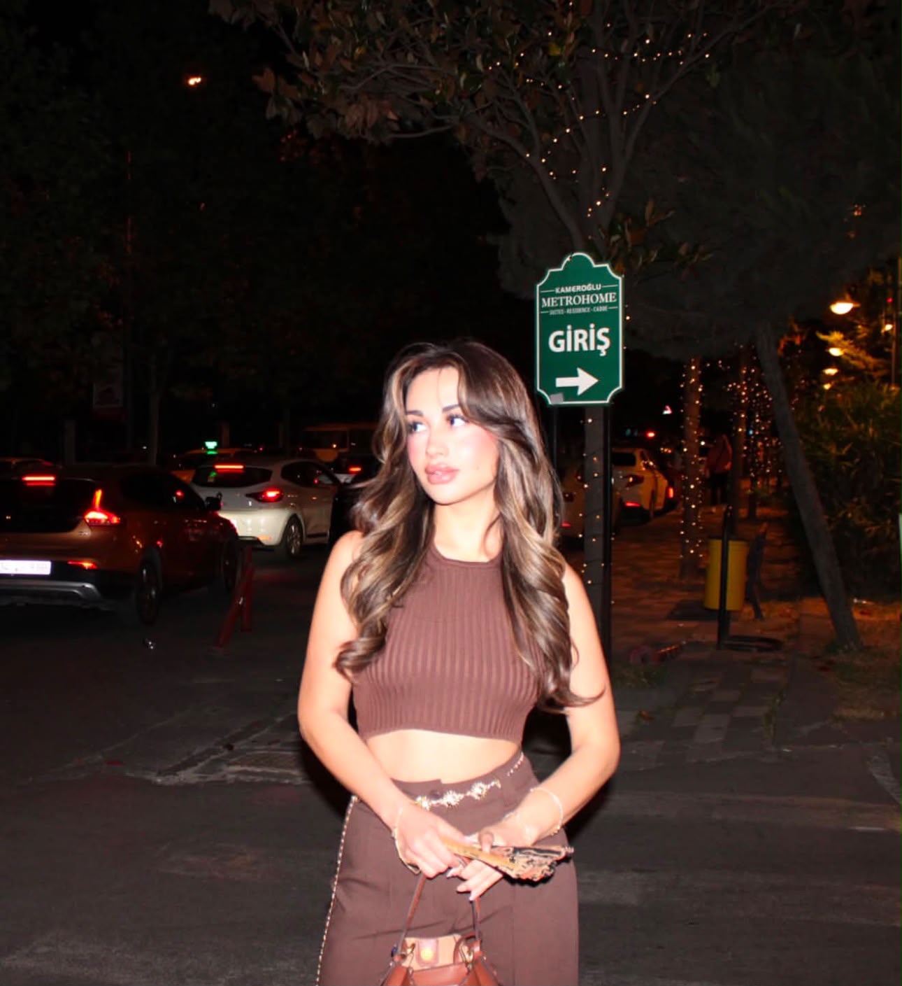Young woman standing on a curb at night, wearing a brown two-piece outfit and holding a small purse, with a green 'Giriş' sign in the background.