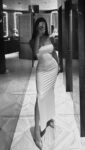 Woman in a tight, strapless white gown with a thigh-high slit posing in a full-length mirror selfie in a stylish bathroom with geometric tiled floor.