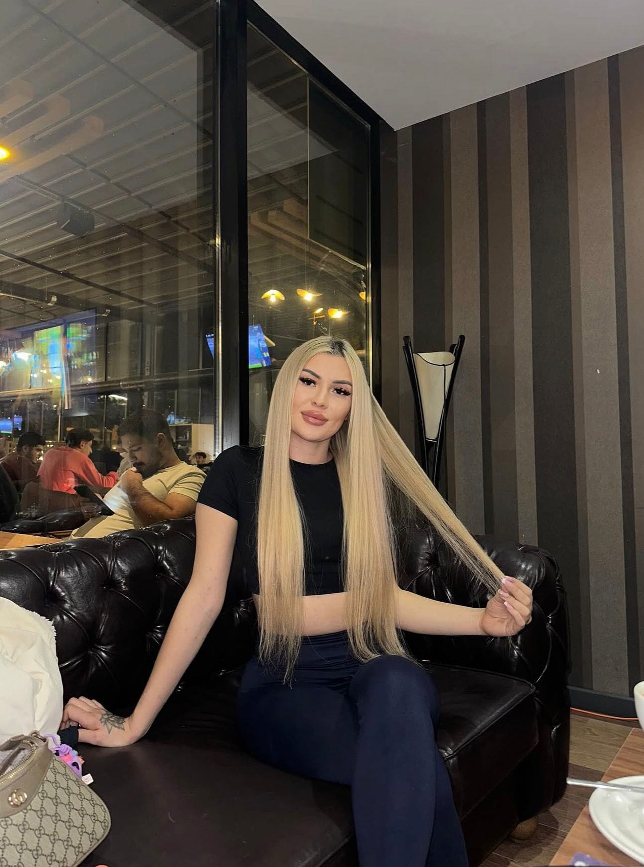 Woman with long, straight blond hair sitting on a black leather couch in a cafe, holding her hair toward camera.