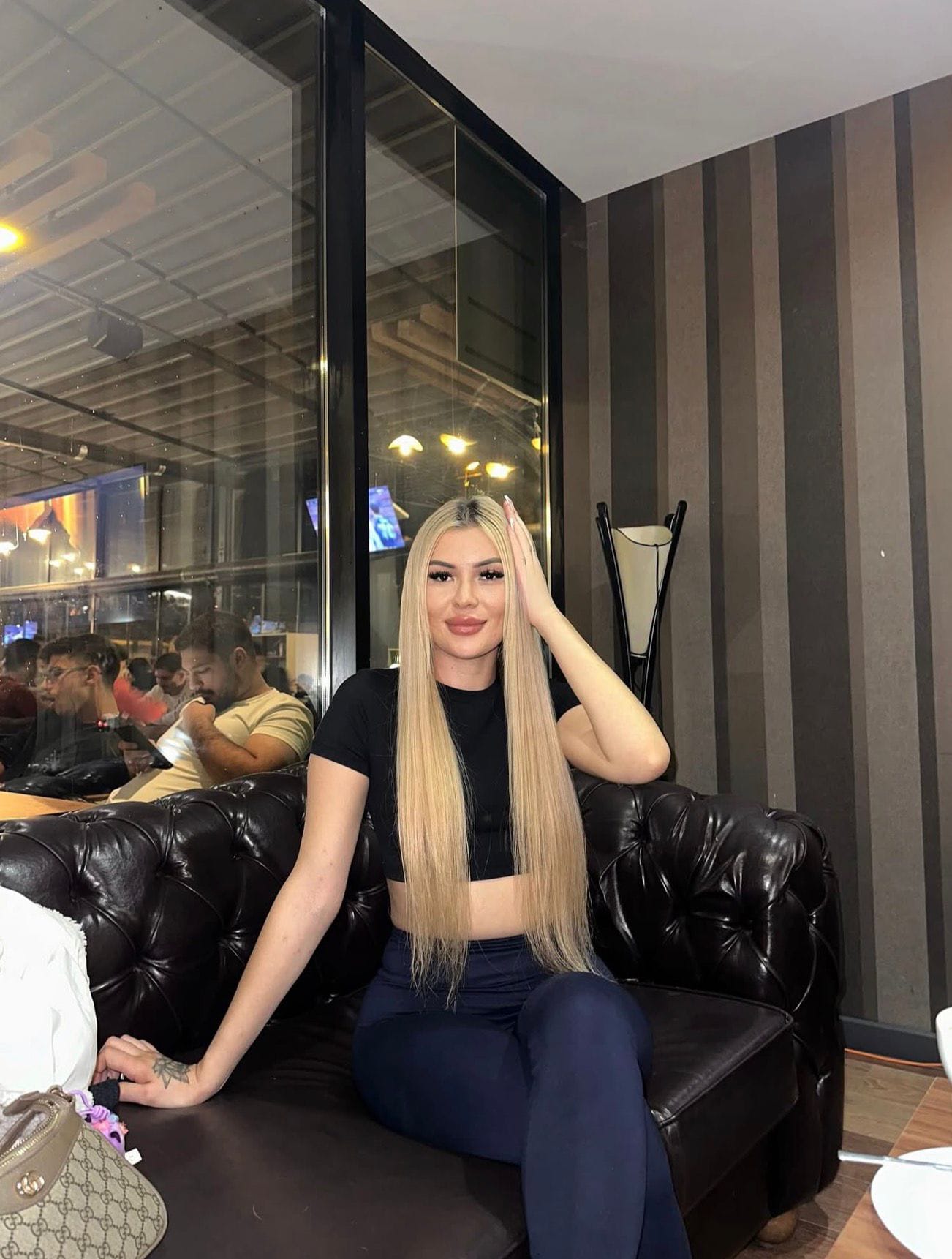 Blonde woman in a black top and navy pants sits on a dark leather couch in a modern lounge with striped walls and glass partition behind her