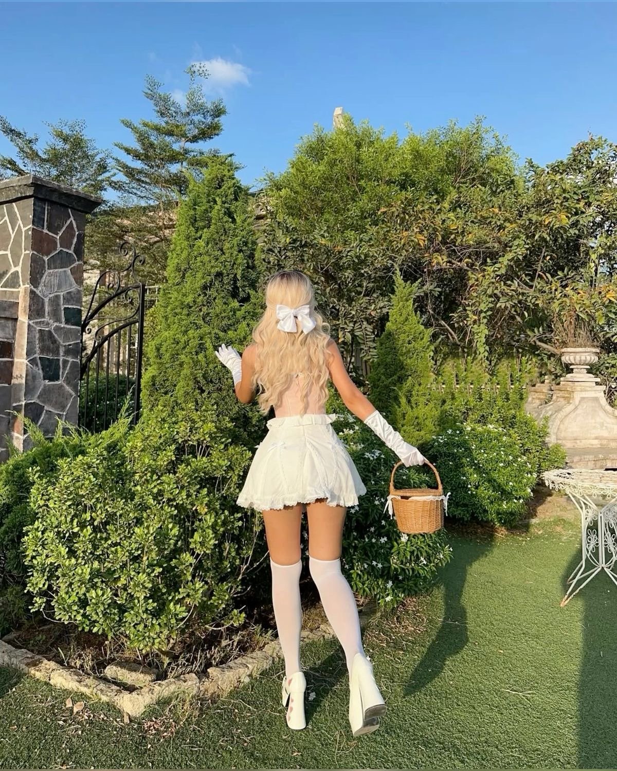Back view of a person in a white cosplay/maid outfit with gloves and a large bow in their blonde hair, carrying a wicker basket in a garden.