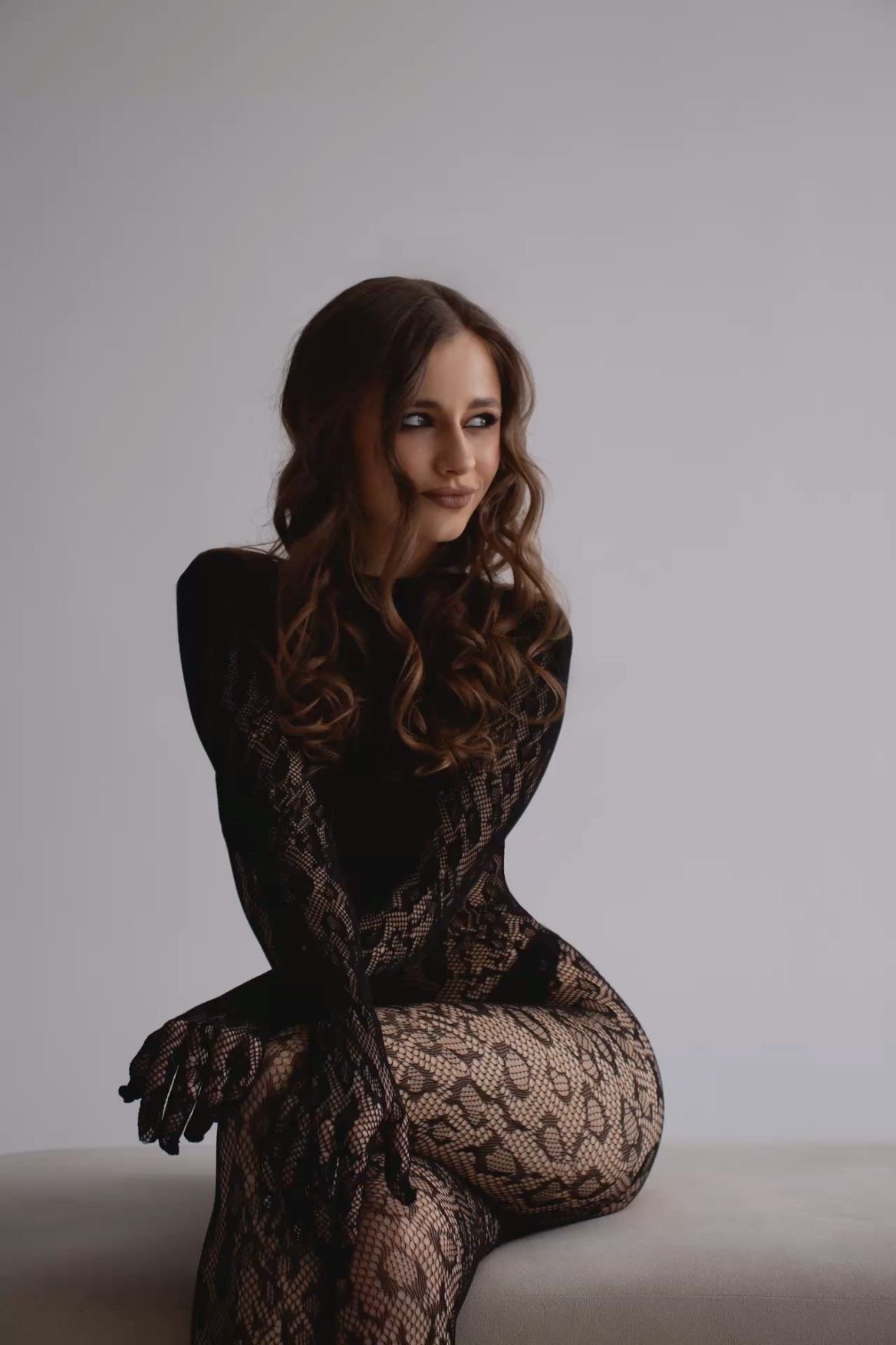 Woman in a black lace bodysuit seated on a light gray backdrop, looking to her left with a soft smile.