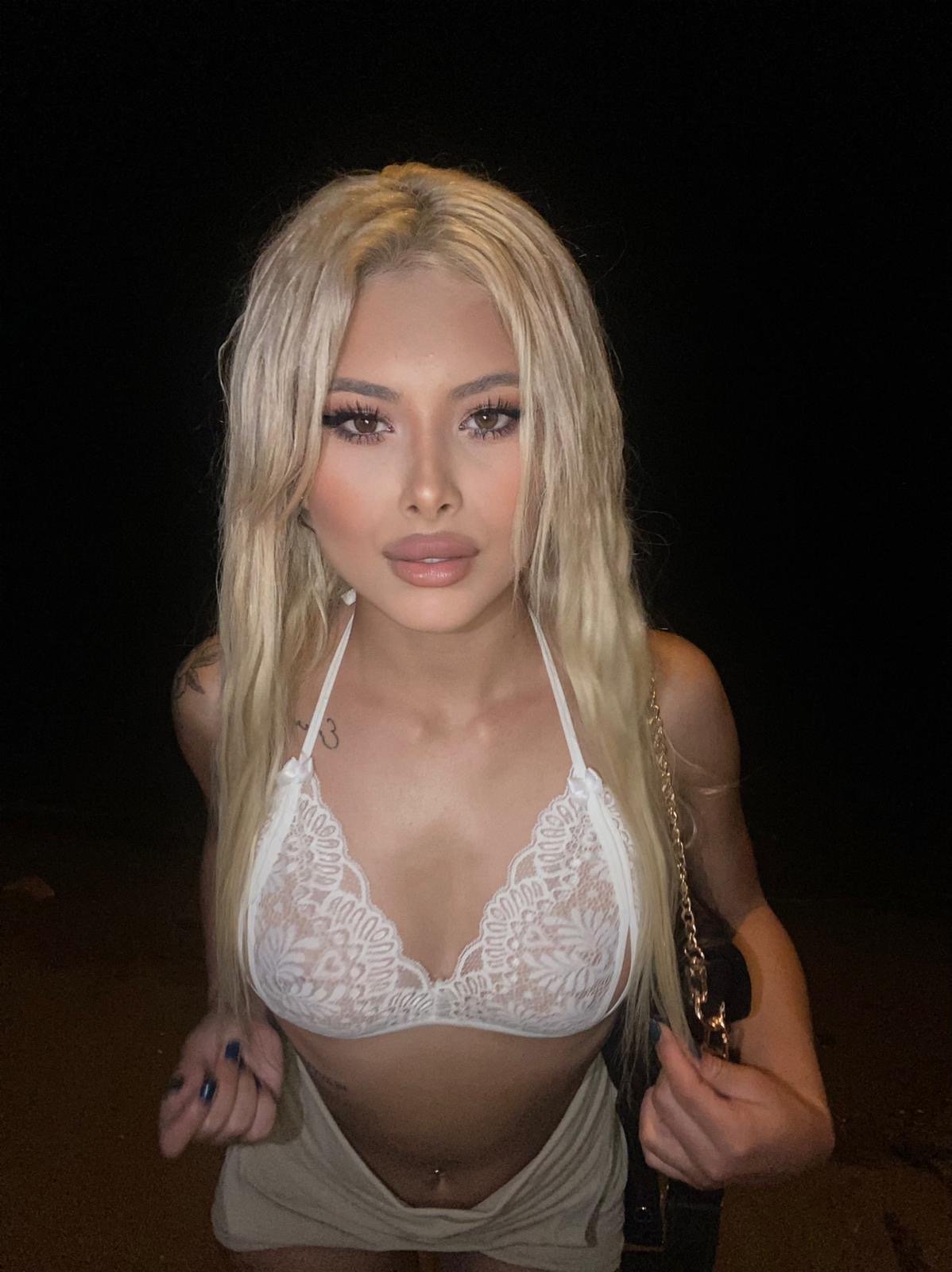 Portrait of a blonde woman in a white lace bralette and light shorts at night, holding a chain-strap bag.