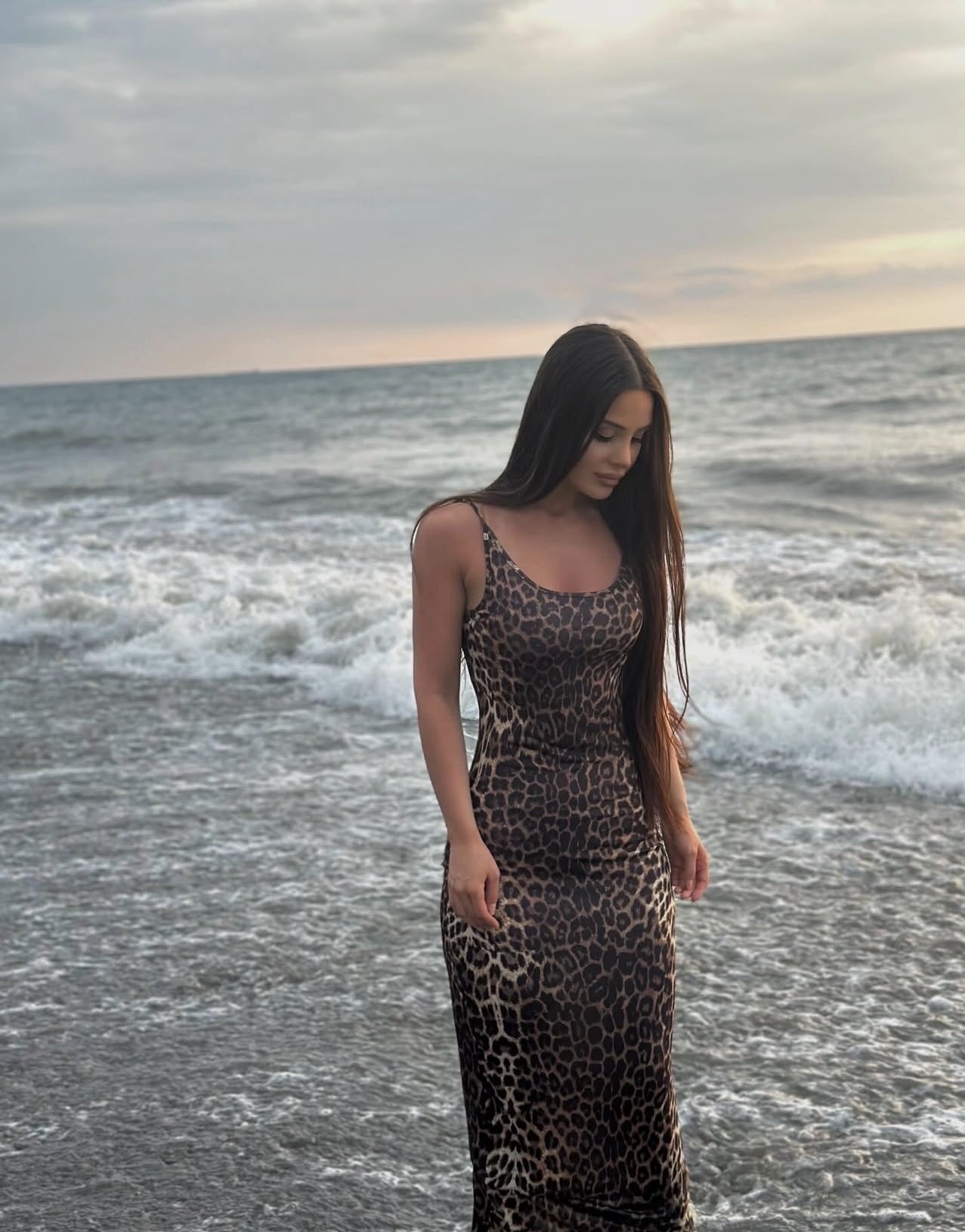 Woman in a leopard-print sleeveless dress standing at the shore, looking down as waves roll in nearby.