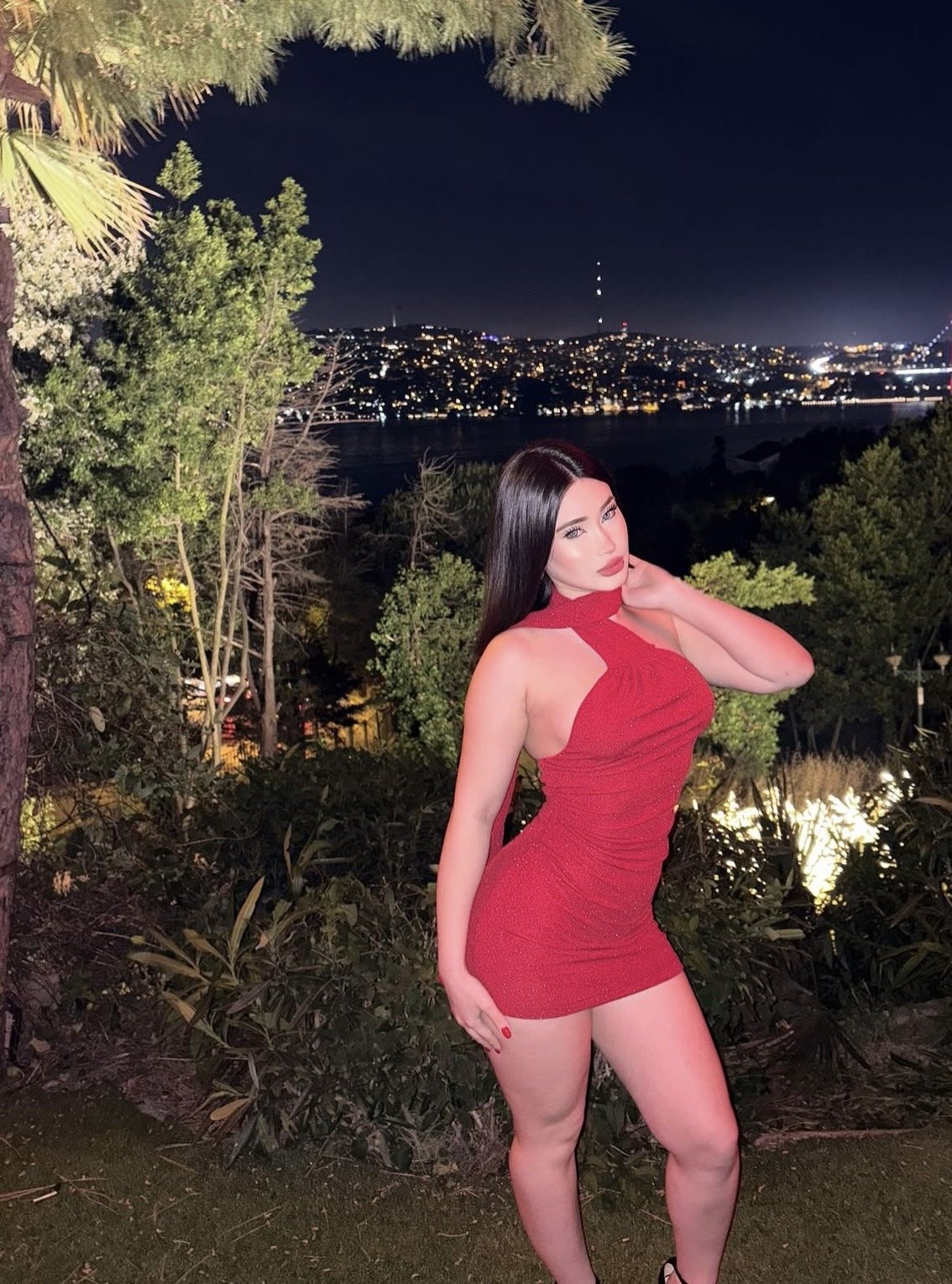 Woman in a red halter mini dress posing outdoors at night, city lights in the distance behind her.