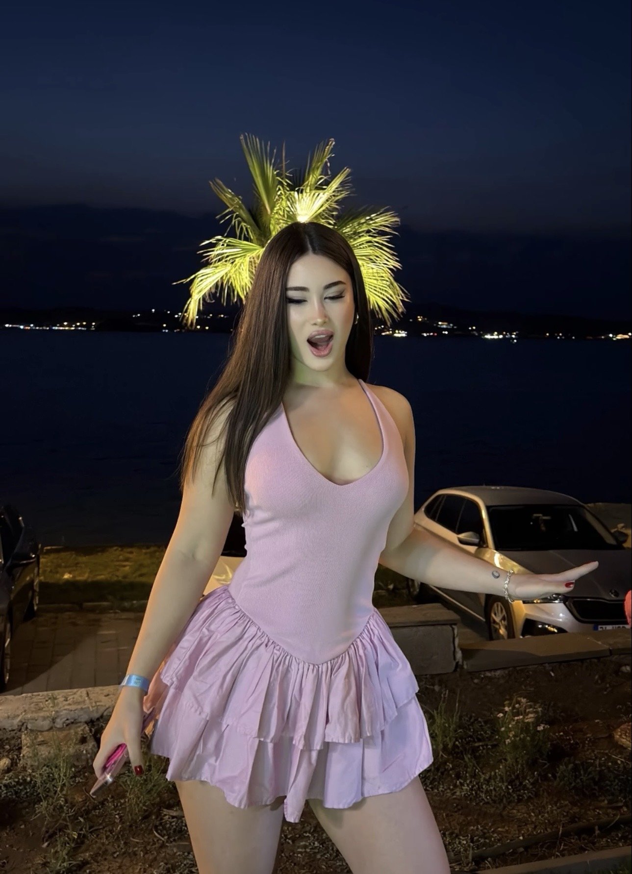 Young woman in a pink sleeveless ruffled dress posing by a waterfront at night, mouth open as if singing, with a lit palm tree behind her.