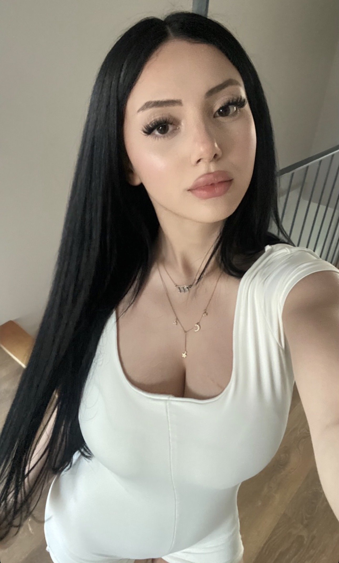 Selfie of a woman with long black hair wearing a white V-neck top and layered gold necklaces, looking at the camera.