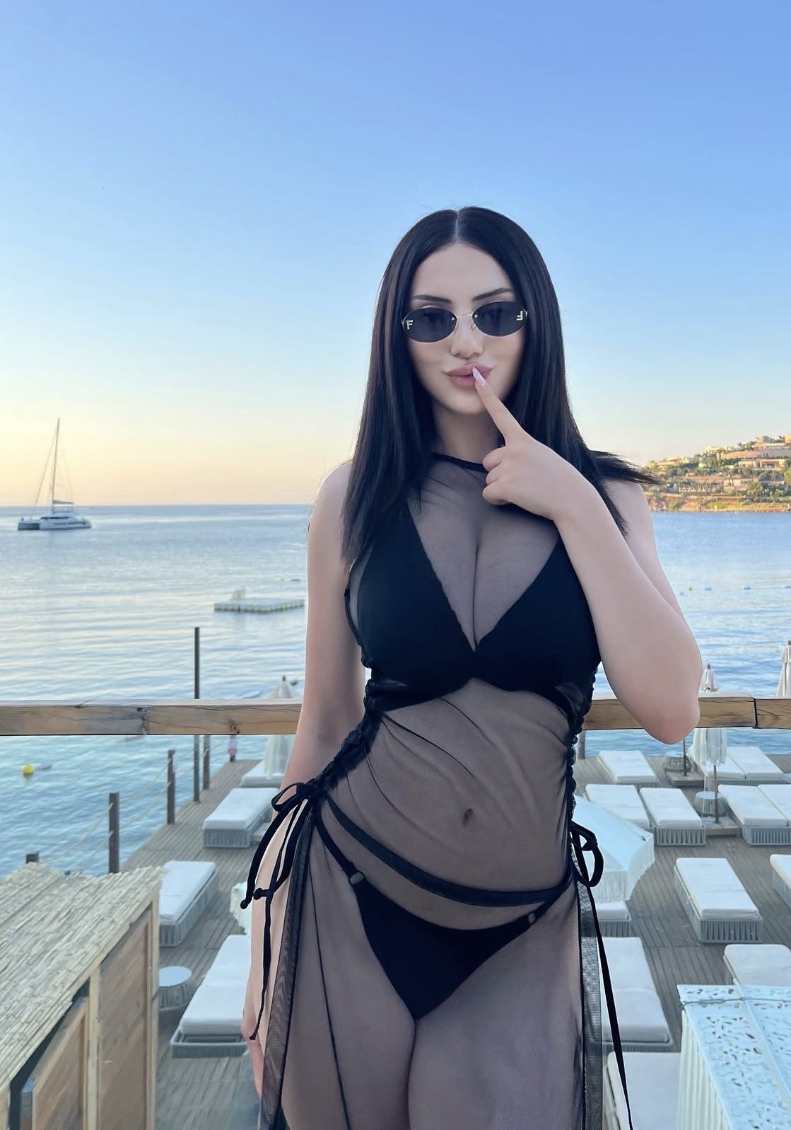 Woman in a black bikini with a sheer black cover-up posing on a wooden deck by calm sea with a boat in the distance, sunny sky.