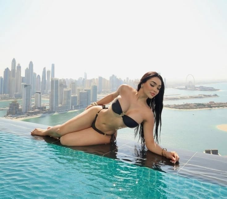 Woman in a black bikini lounging on the edge of an infinity pool, with a city skyline (Dubai) and a large Ferris wheel in the background.