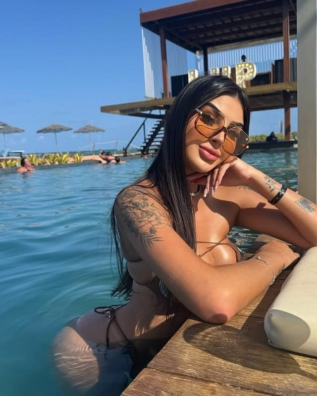 Woman in a bikini leaning on poolside railing, wearing sunglasses, with tattoos visible, sunny resort scene in the background.