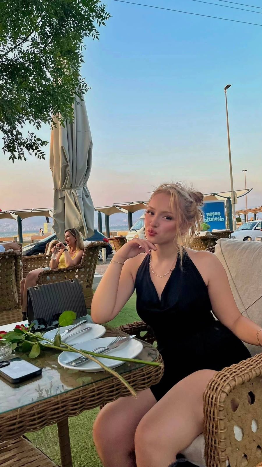 Woman in a black halter dress sits at a wicker table by the beach, blowing a kiss toward the camera with a rose on the plate in front.