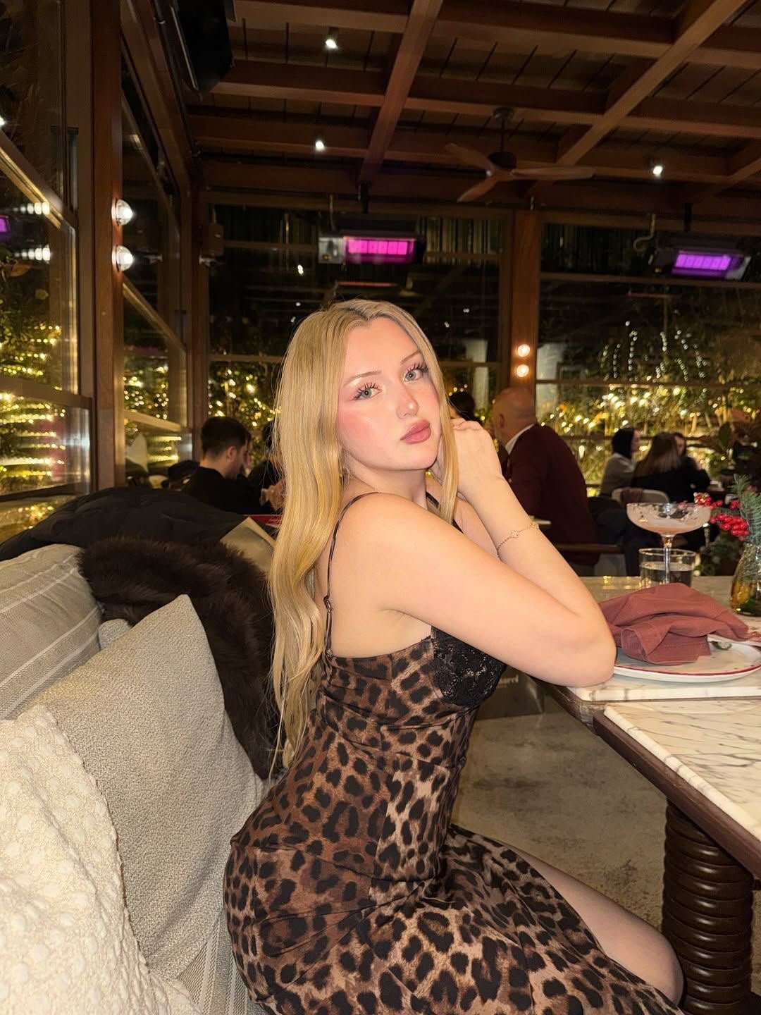 Woman with long blonde hair in a leopard print dress seated at a restaurant table, posing for the camera.