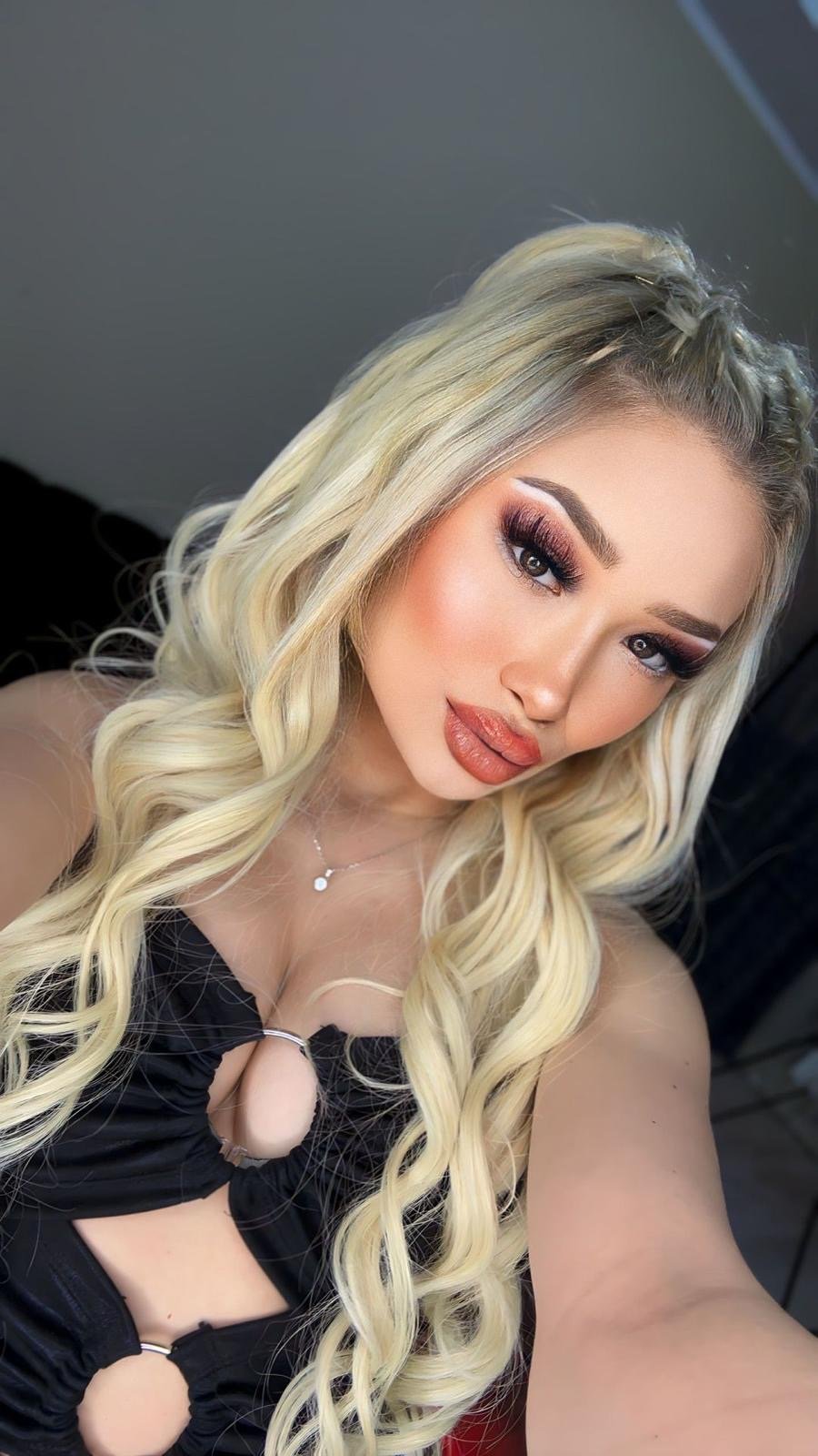 Close-up selfie of a woman with long blonde curls and dramatic makeup, wearing a black cut-out top.