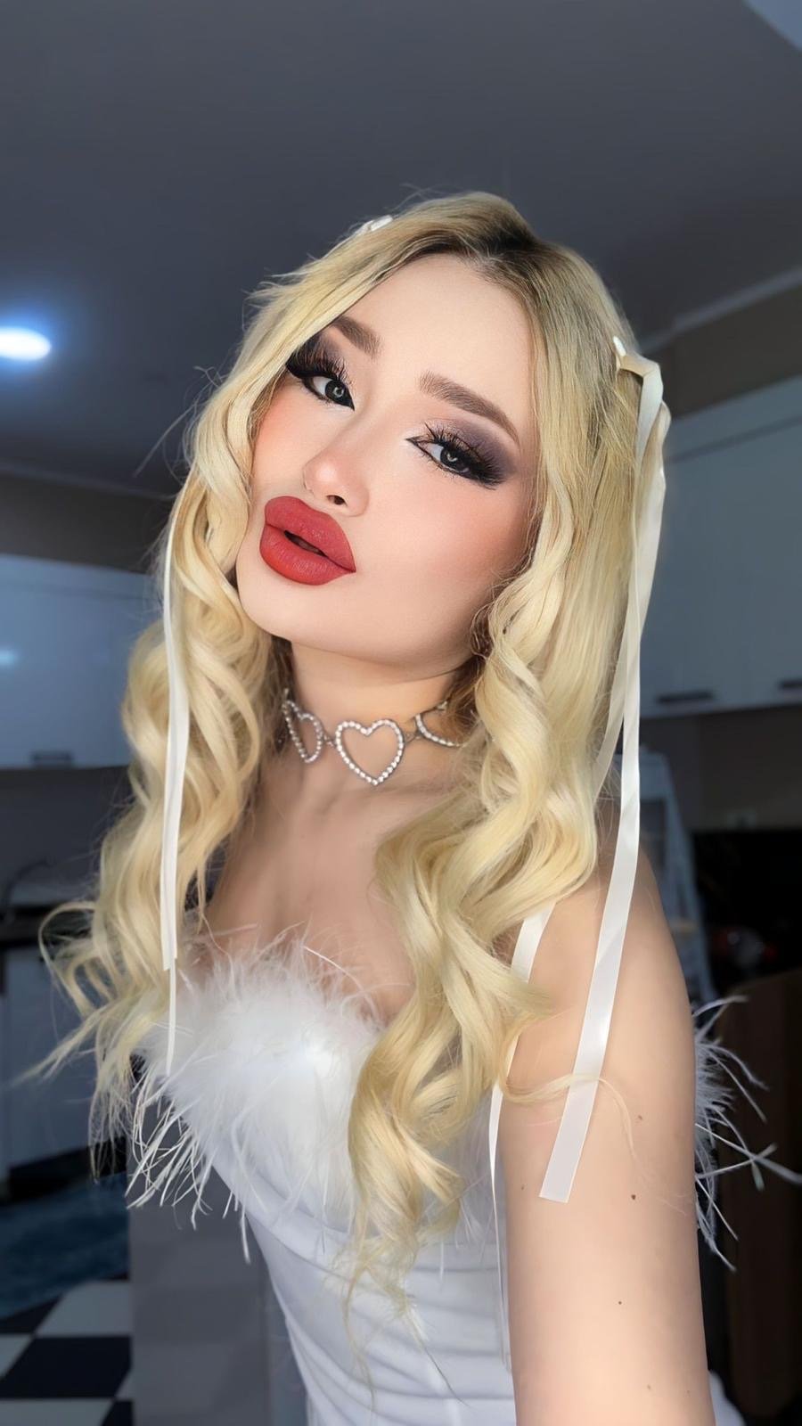 Close-up portrait of a person with long blonde curls, dramatic smoky eye makeup and bold red lips, wearing a pearl heart choker against a white outfit.