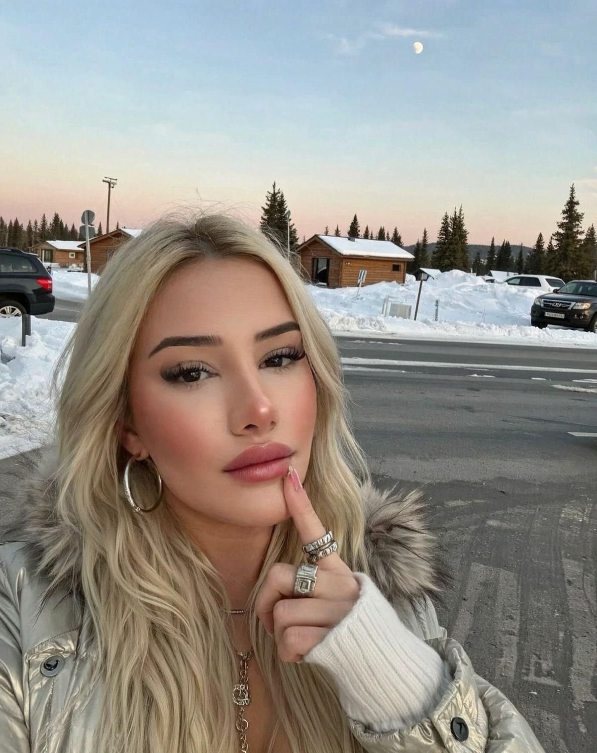 Close-up selfie of a blonde woman wearing a silver fur-lined jacket, large hoop earrings, and multiple rings, posing with her finger on her lips in a snowy parking area with cabin buildings in the background.