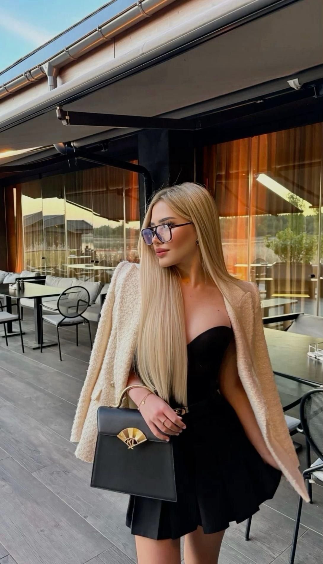Stylish woman with long blonde hair and glasses posing outside a cafe in a black strapless dress with a beige coat, holding a black handbag.