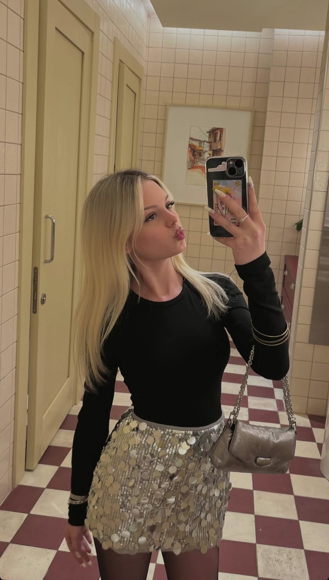 Woman in a black long-sleeve top and gold sequin skirt taking a mirror selfie in a tiled bathroom, puckering lips.