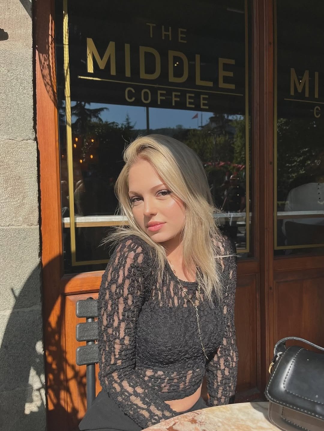 Woman with blonde hair in a black lace top sits at an outdoor café table, sunlight on her face with 'The Middle Coffee' sign in the window behind her.