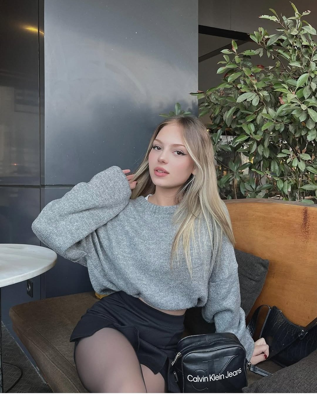 Young woman with blonde hair sits on a wooden bench at an outdoor cafe, wearing a gray knit sweater and a black skirt, with a black Calvin Klein bag nearby.
