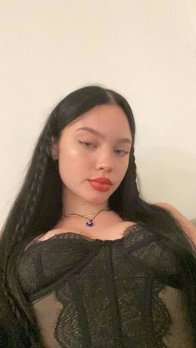 Young woman with long black hair wearing a black lace top and a blue pendant necklace, posing against a light wall.