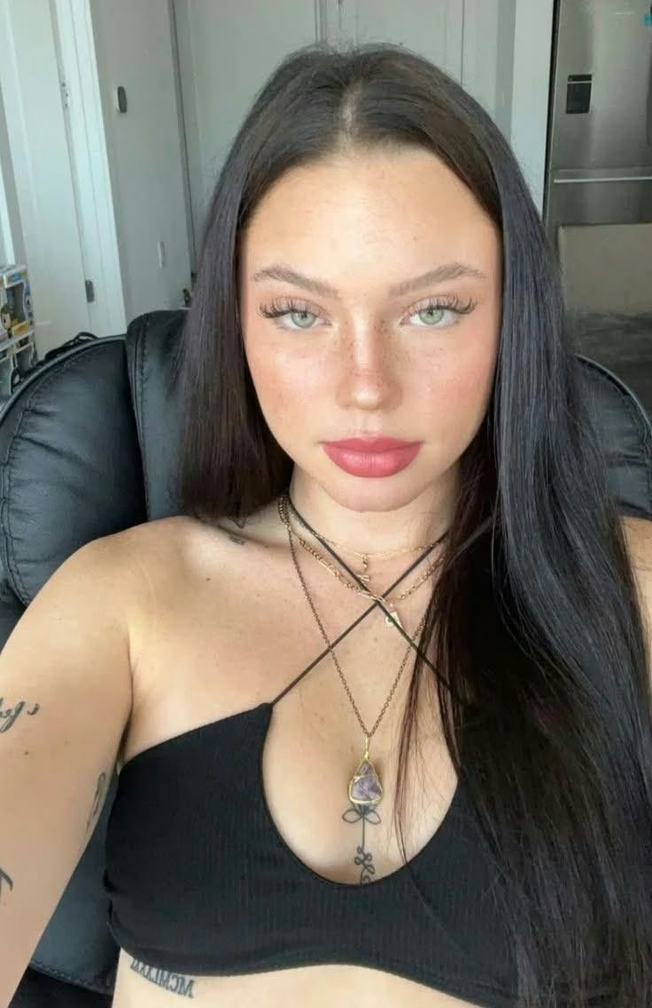 Portrait of a young woman with long dark hair wearing a black halter top, sitting in a chair and looking at the camera, wearing layered necklaces and a pendant.