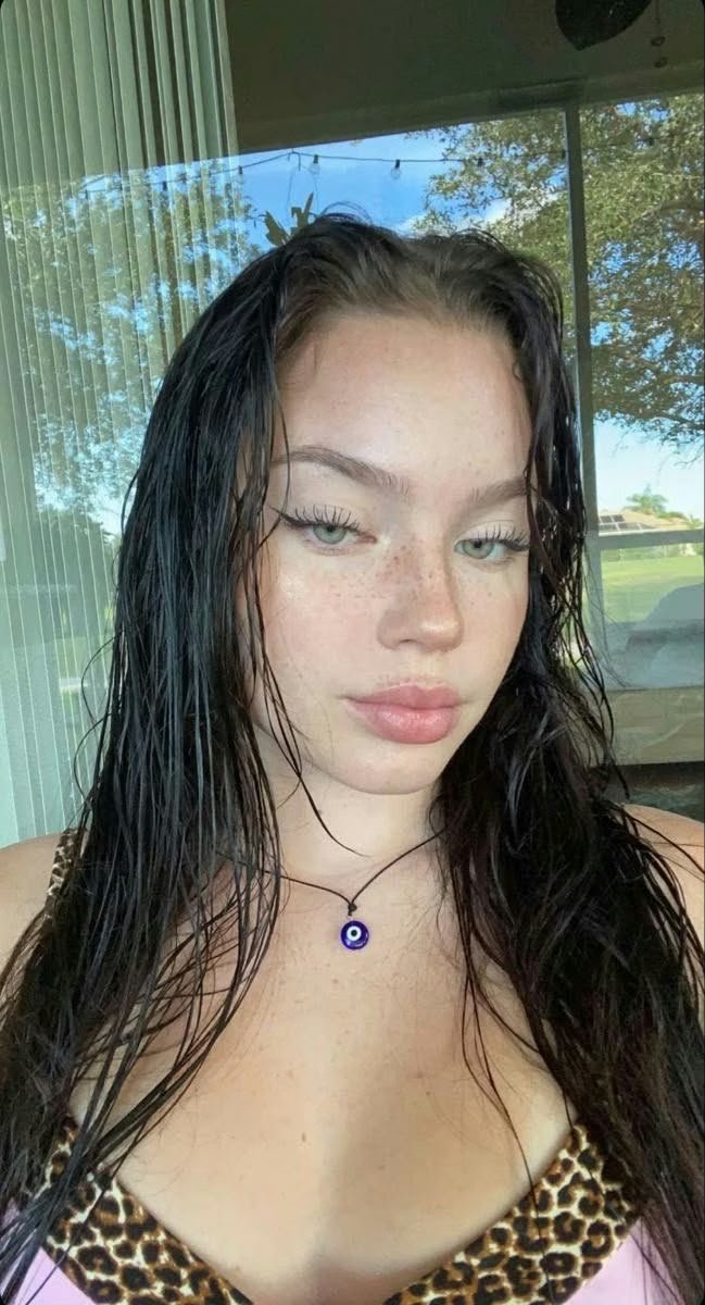 Close-up of a young woman with wet dark hair, wearing a leopard-print top and a blue evil-eye necklace outdoors.
