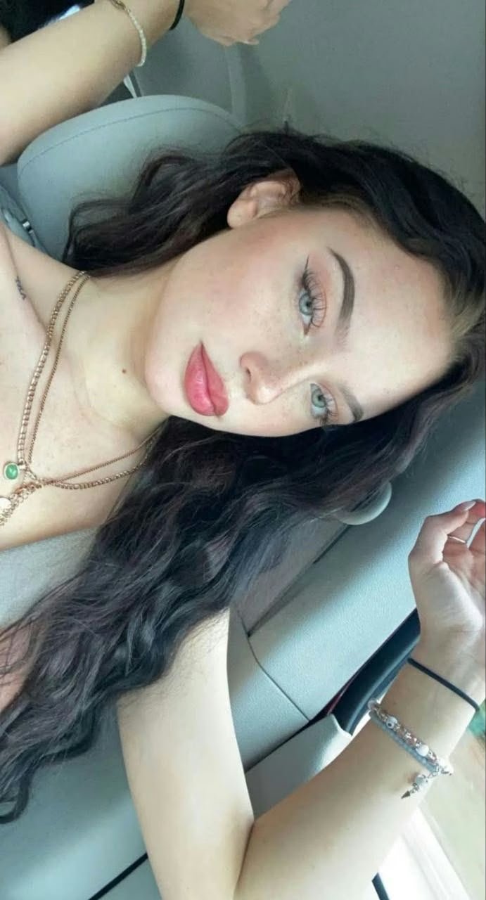 Young woman in a car with long dark hair, pink lipstick, blue eyes, wearing layered necklaces and bracelets.