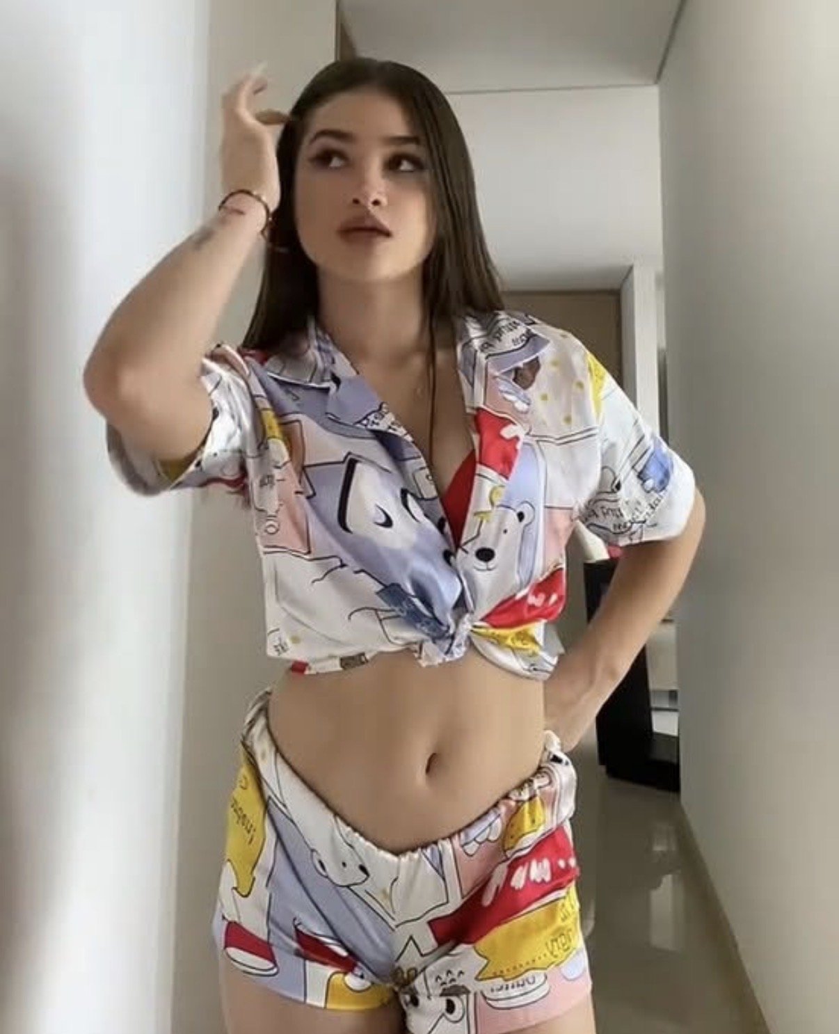 Woman in a colorful cartoon-print two-piece outfit, knotting the shirt to show midriff, posing with hand in hair in a bright hallway.