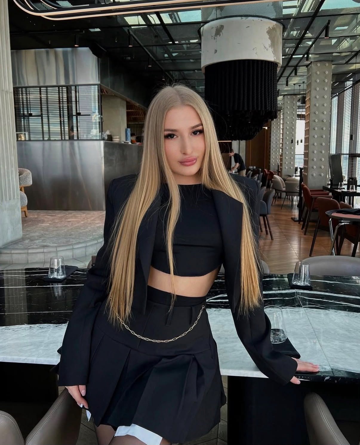 Young woman with long blonde hair in a black blazer and crop top posing at a stylish restaurant table.