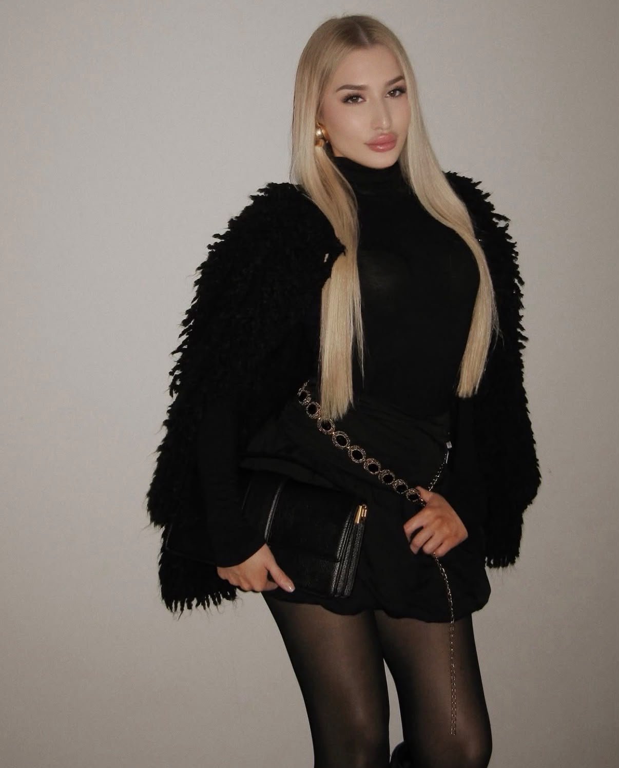 Woman with long blonde hair wearing a black turtleneck and a fuzzy black coat, holding a small black purse with a chain strap.