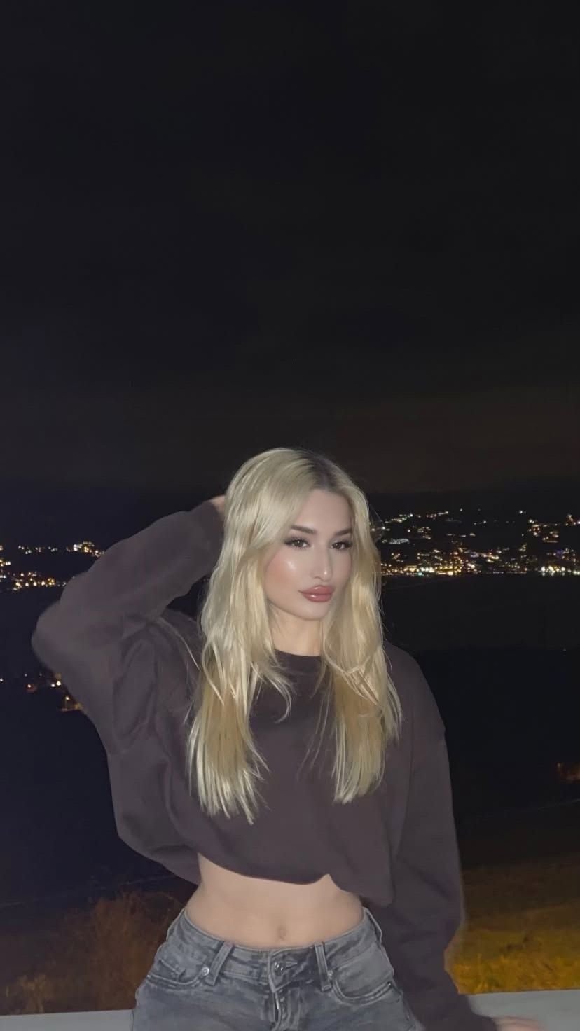 Woman with long blonde hair posing at night, wearing a cropped dark hoodie and jeans against a city-lit background.