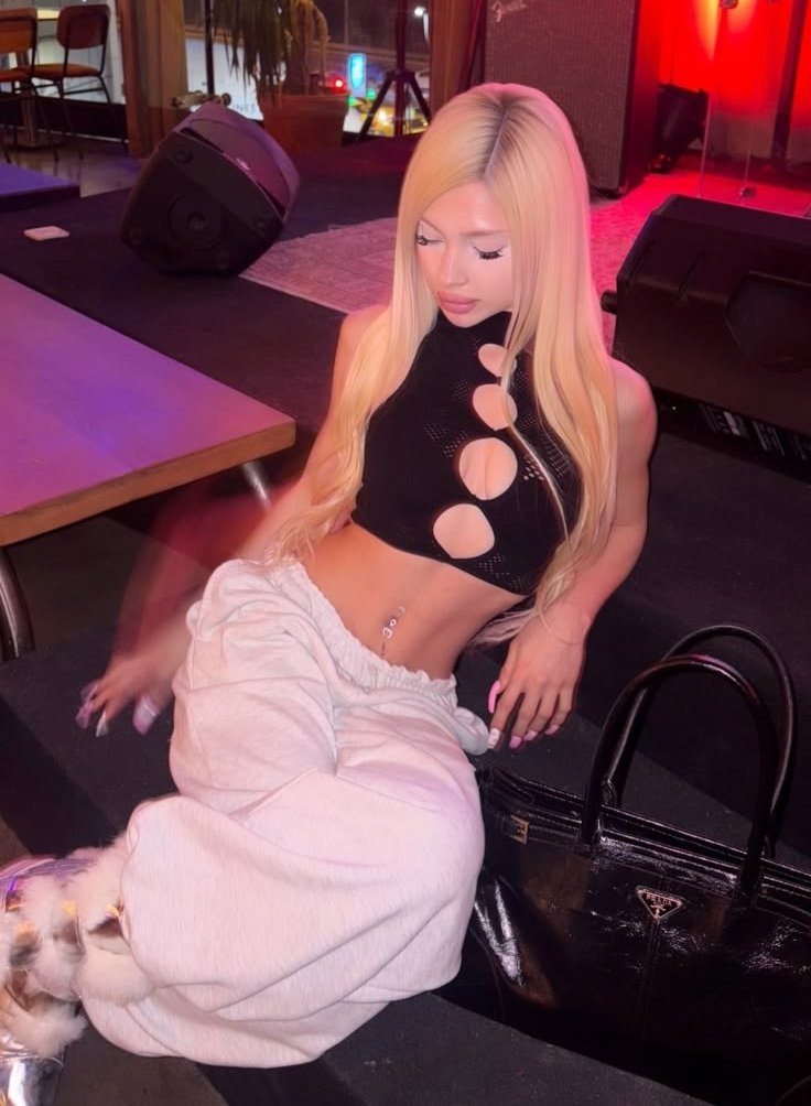 Young woman with long blonde hair seated on a stage floor in a black cut-out crop top and white sweatpants, beside a black handbag.