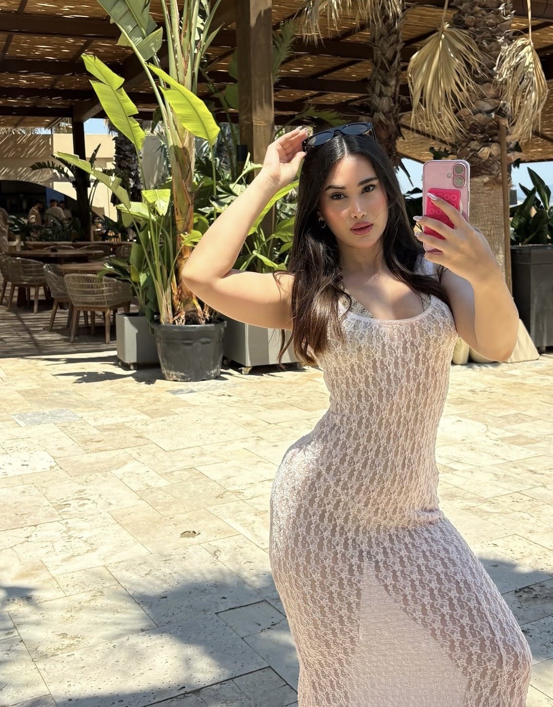 Woman in a beige lace dress takes a selfie outdoors at a tropical cafe, sunglasses resting on her head.