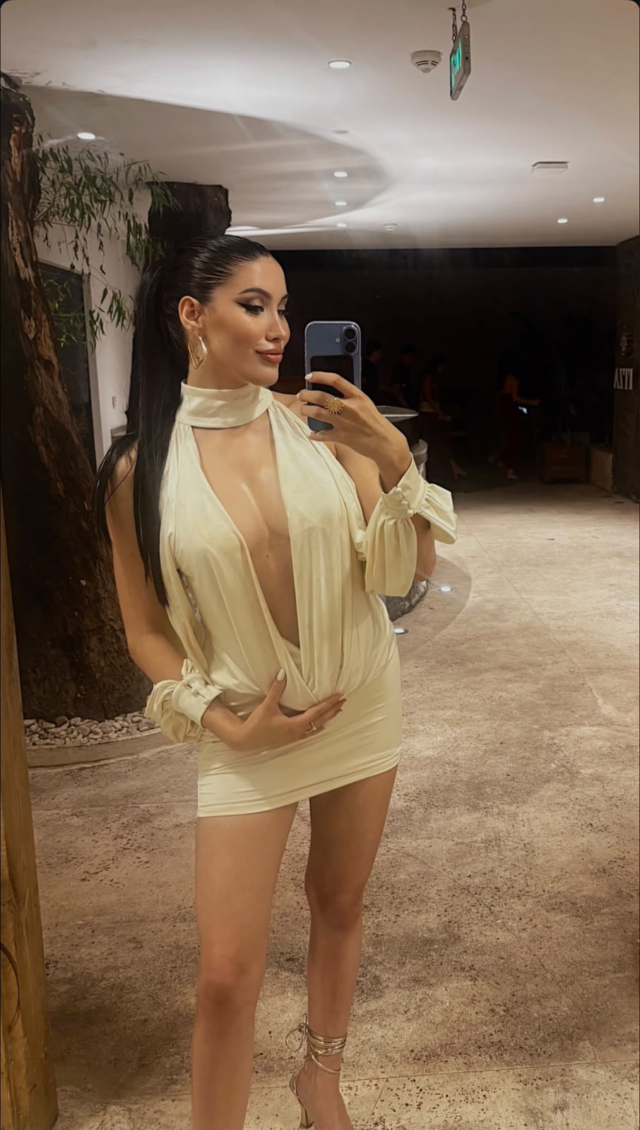 Woman taking a mirror selfie in a cream halter mini dress with a deep V neckline, gold hoop earrings, and strappy heels.