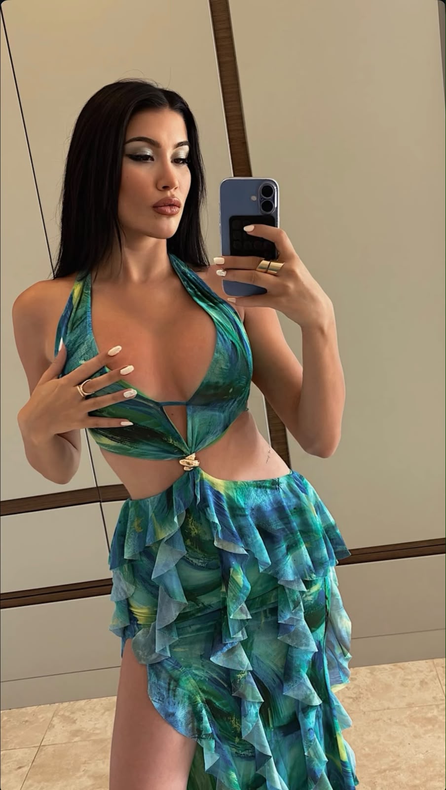 Woman in a green-blue halter bikini with a layered ruffle skirt posing for a mirror selfie, holding a blue smartphone.