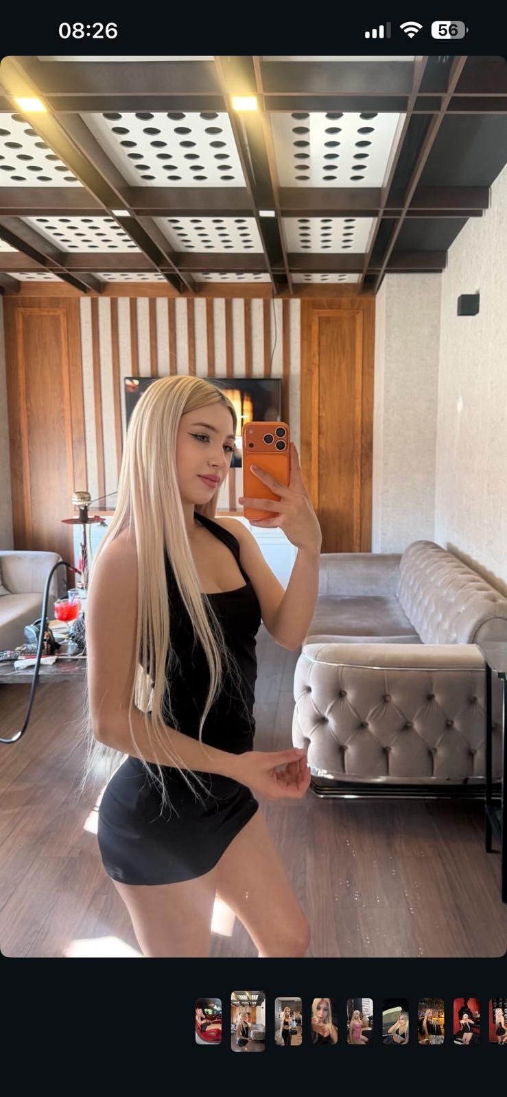 Woman with long blonde hair in a black sleeveless dress takes a mirror selfie in a stylish lounge with a tufted sofa.