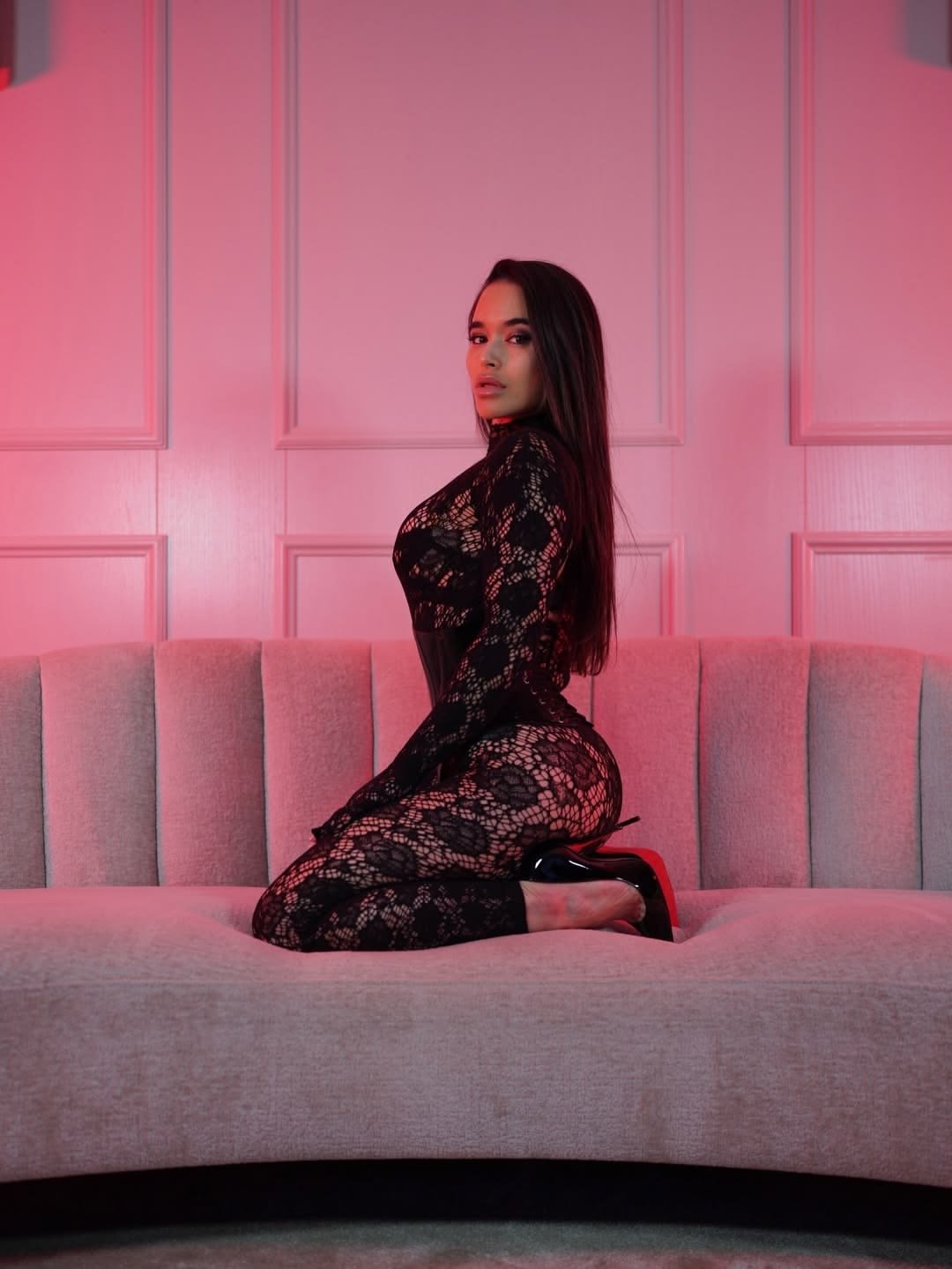 Woman kneeling on a light gray sofa wearing a black lace bodysuit and redHigh heels, bathed in pink lighting, facing sideways.