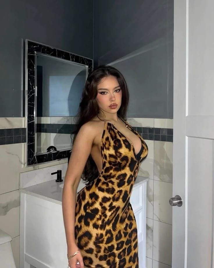 Woman in a leopard-print halter dress standing in a bathroom, posing for the camera by a sink and mirror.