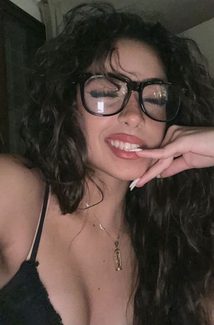 Young woman with long curly hair wearing oversized black glasses, a black strappy top, and a gold pendant necklace, smiling with her hand near her mouth.