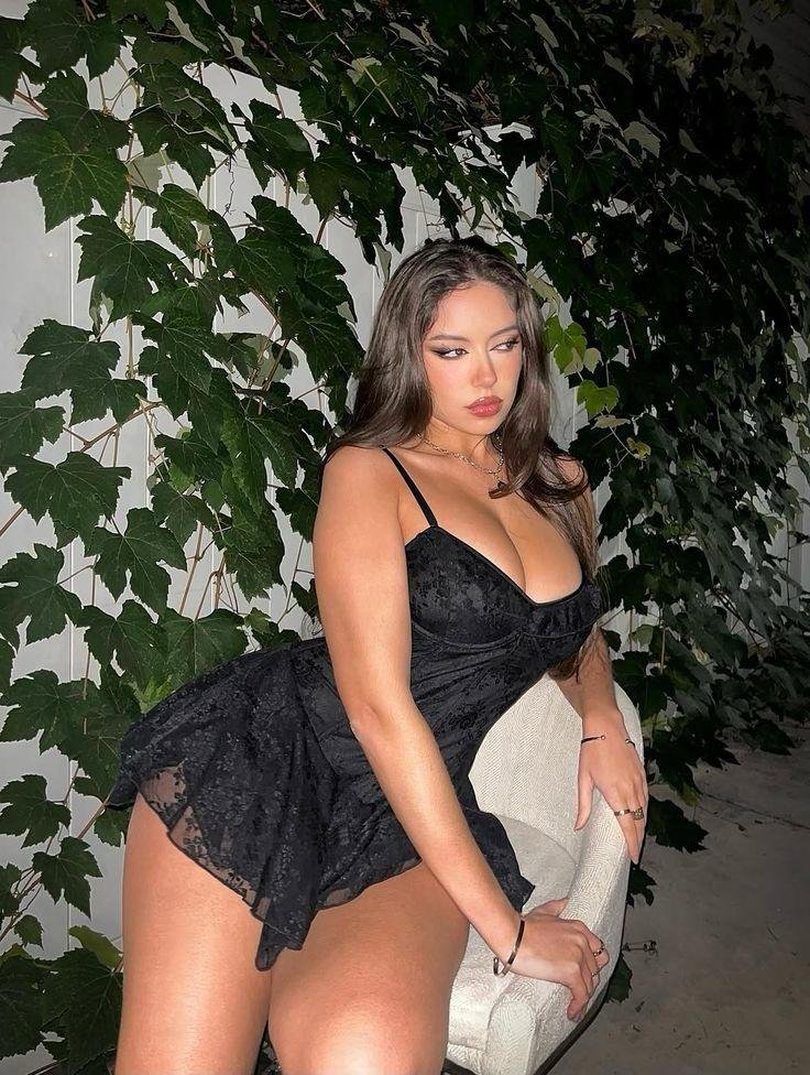 Woman in a black lace dress posing on a chair in front of a wall covered with ivy vines behind her.