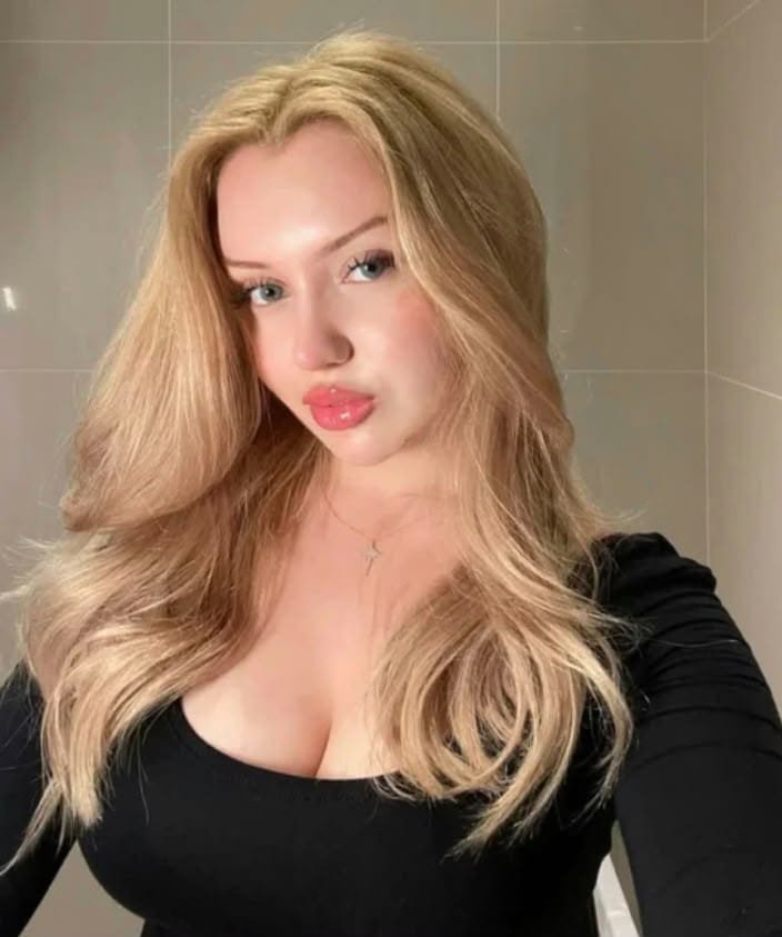 Self-portrait of a blonde woman with long wavy hair wearing a black top, standing in a tiled bathroom and looking at the camera.