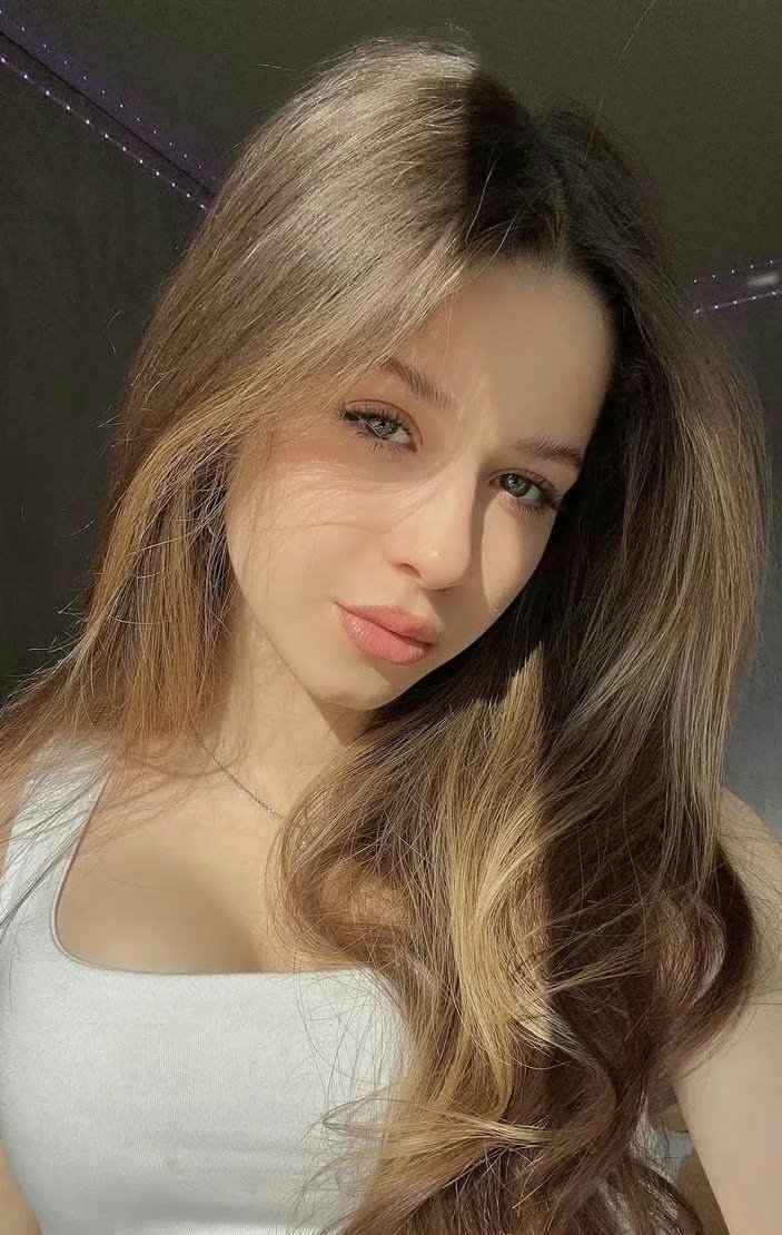 Young woman with long light brown hair, sunlit face, wearing a white tank top and a subtle smile.