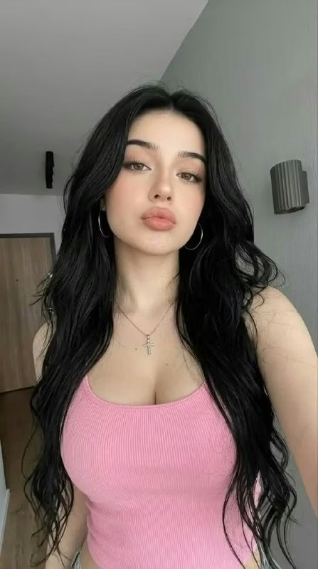 Portrait of a young woman with long black wavy hair, pink ribbed tank top, hoop earrings, and a cross pendant necklace indoors.