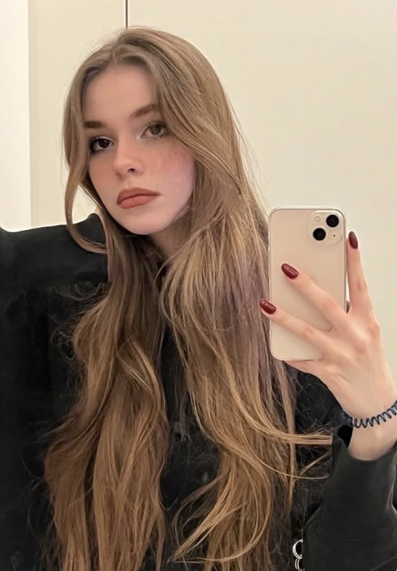 Woman with long, wavy light brown hair taking a mirror selfie with a gold iPhone, wearing a black top and burgundy nail polish.