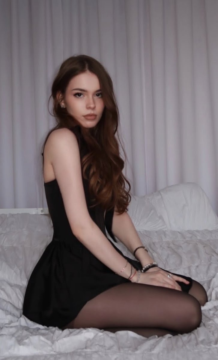 Woman with long brown hair sitting on a white bed in a black sleeveless dress, facing the camera.