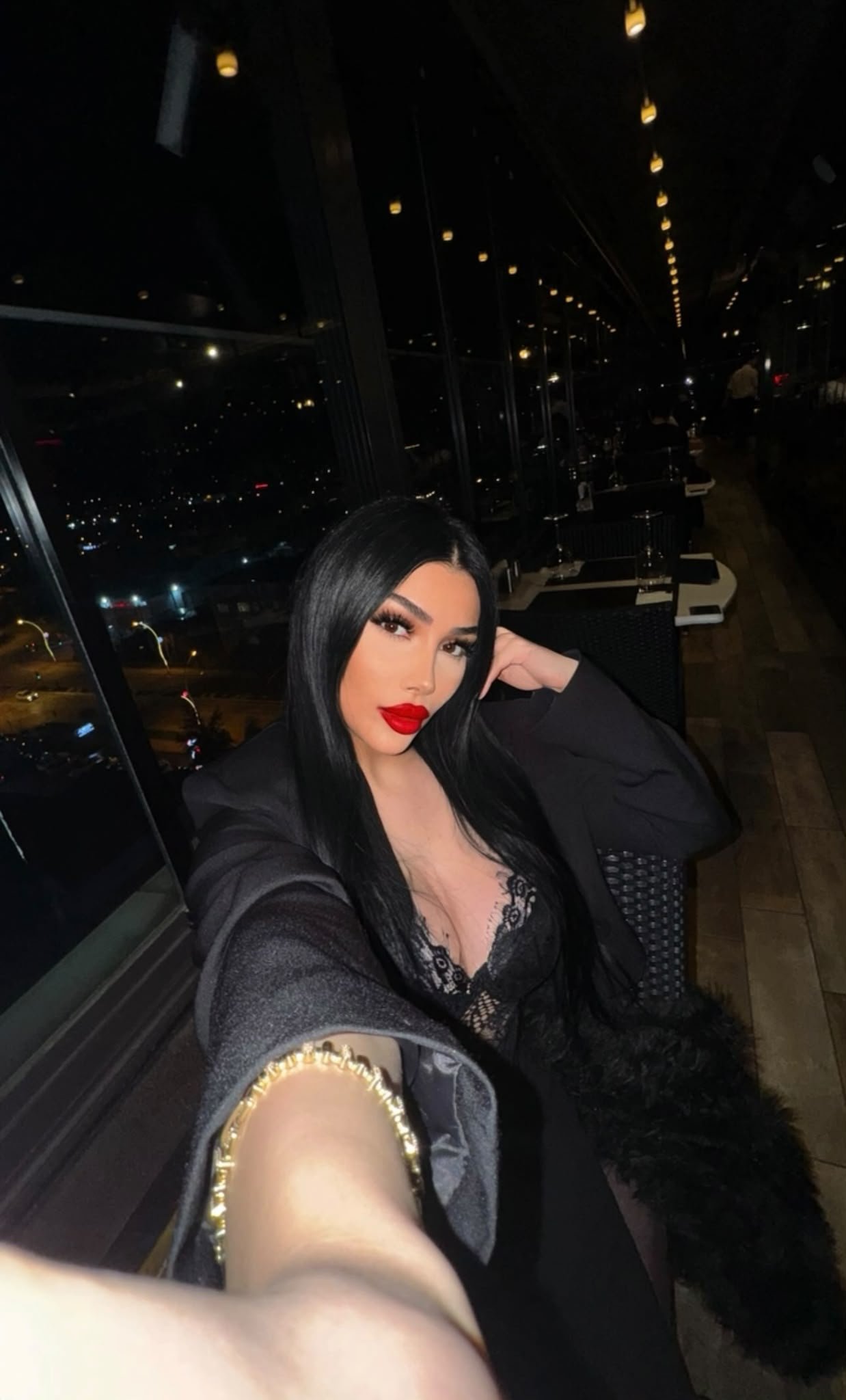 Self-portrait of a woman with long black hair wearing red lipstick and a black lace top, taking a selfie at night in a dim restaurant with city lights outside.