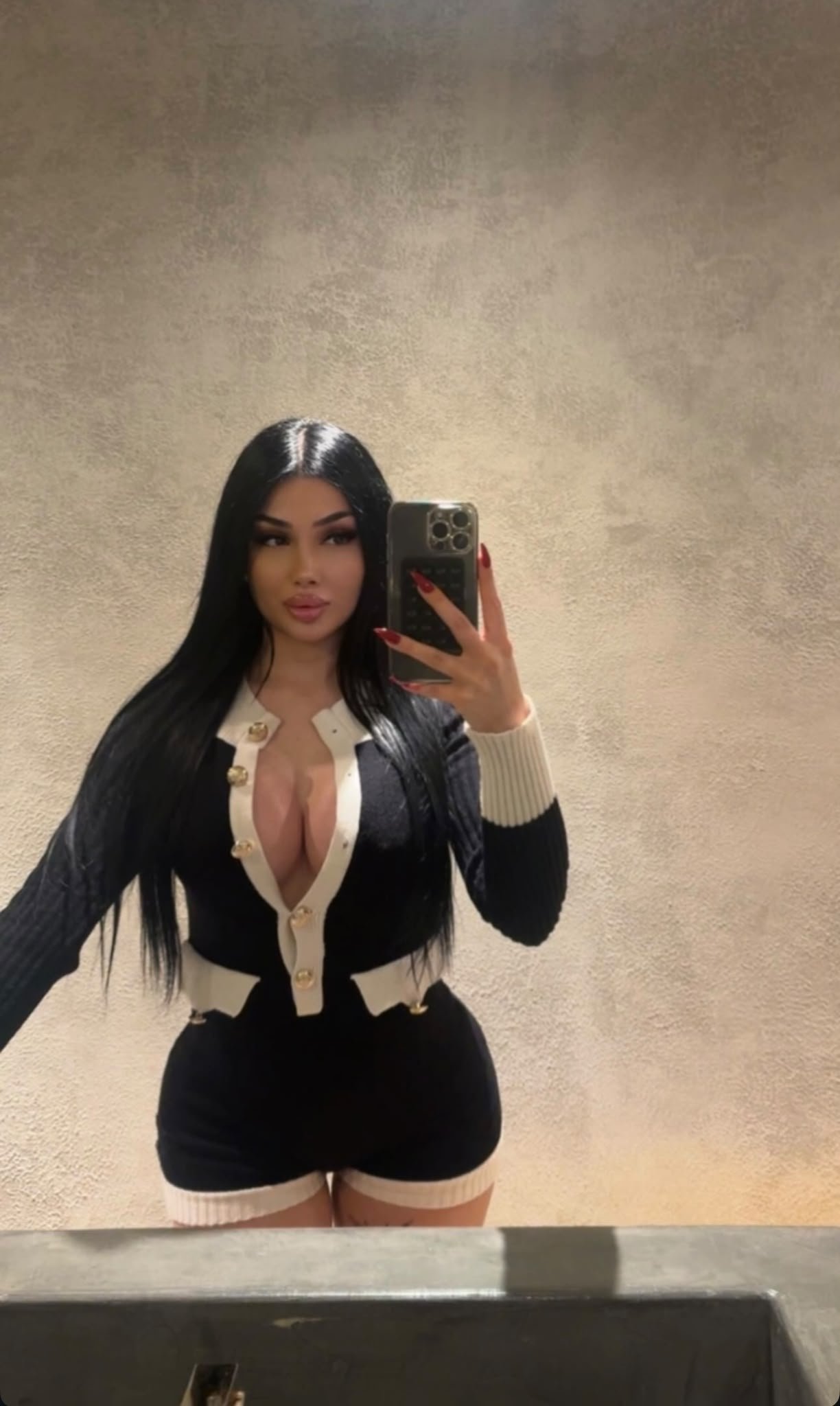 Mirror selfie of a woman with long black hair wearing a black and cream cardigan with gold buttons and a matching black skirt in a beige-textured bathroom wall background, holding a phone with a large camera setup.